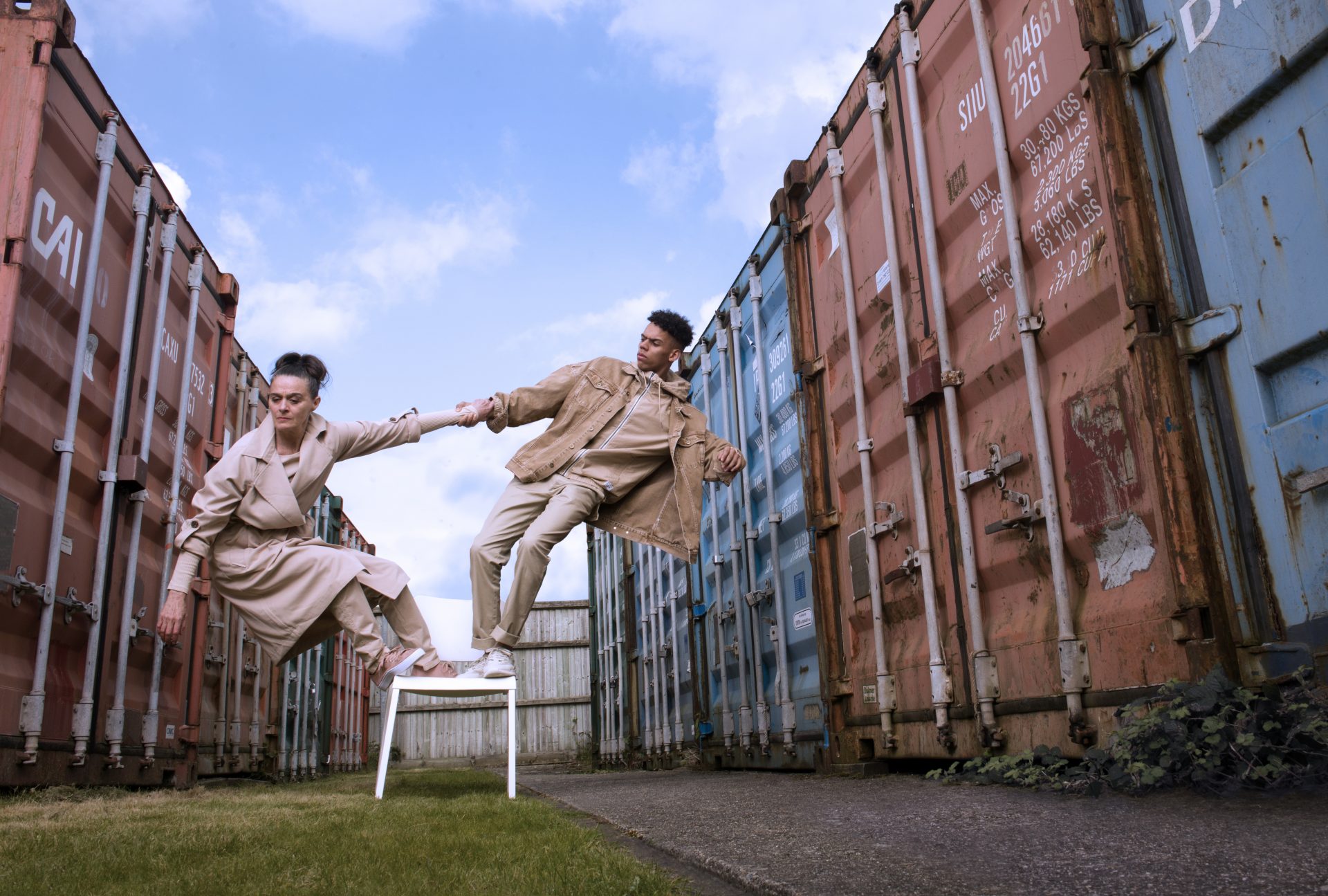 Two dancers hold hands while balancing on a chair. They are surrounded by industrial storage units.