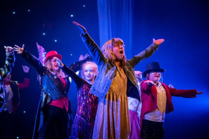 The Remix inclusive performing arts group. Young girl with glasses in foreground wearing a gold dress and denim jacket holding her arms in the air. Three other people behind listing their arms, two are wearing top hats.