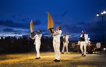 Four men in white suits throw corn in the air.