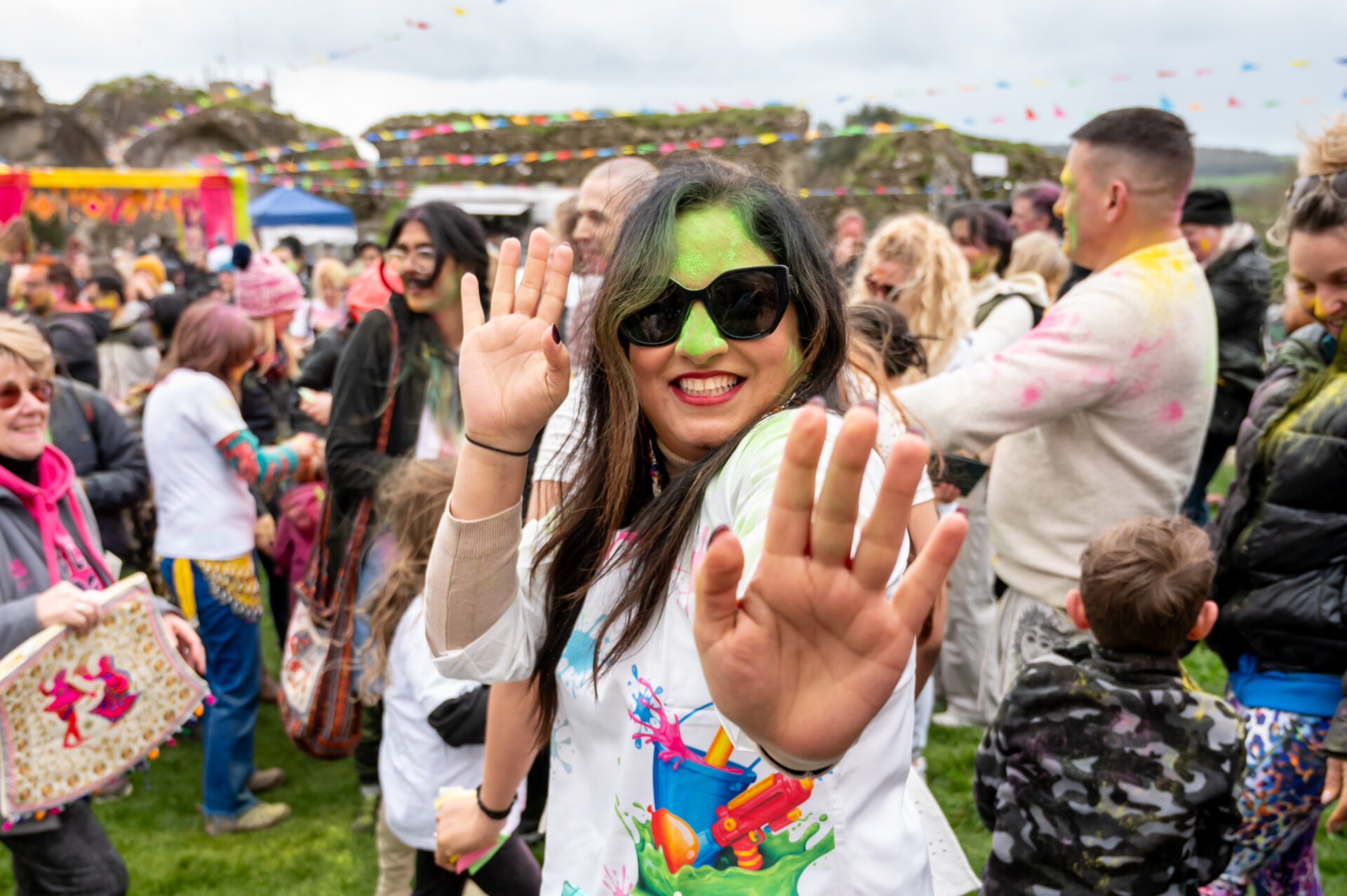 An Indian woman wearing sunglasses with bright green paint on her face, smiles at the camera. A large crowd of people is behind her.