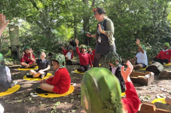 School children sit on the ground in a woods, wearing green helmets. A woman walks amongst them.