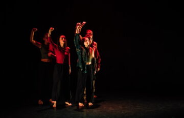 A group of dancers on a dark stage, all holding their right hand in the air.