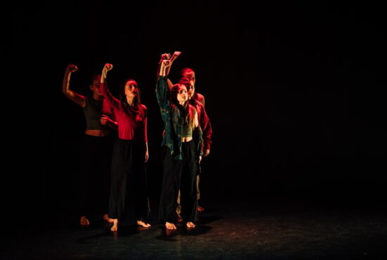 A group of dancers on a dark stage, all holding their right hand in the air.