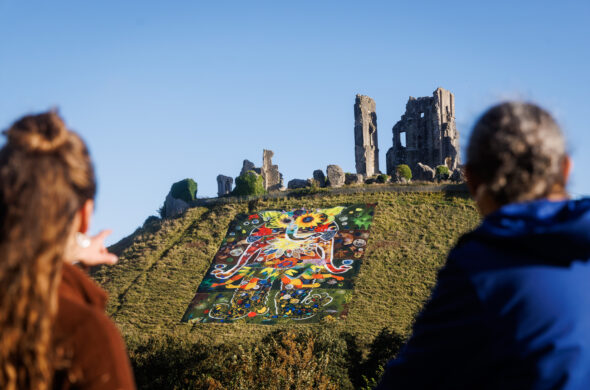 The backs of two people's heads who are looking at a large canvas on a green hill with an ancient castle in the background.