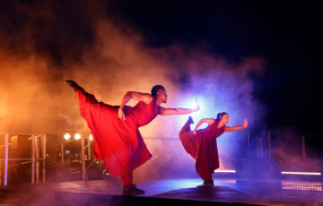 Two dancers with outstretched arms and legs are light from behind. They wear baggy red clothing.