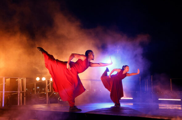 Two dancers with outstretched arms and legs are light from behind. They wear baggy red clothing.