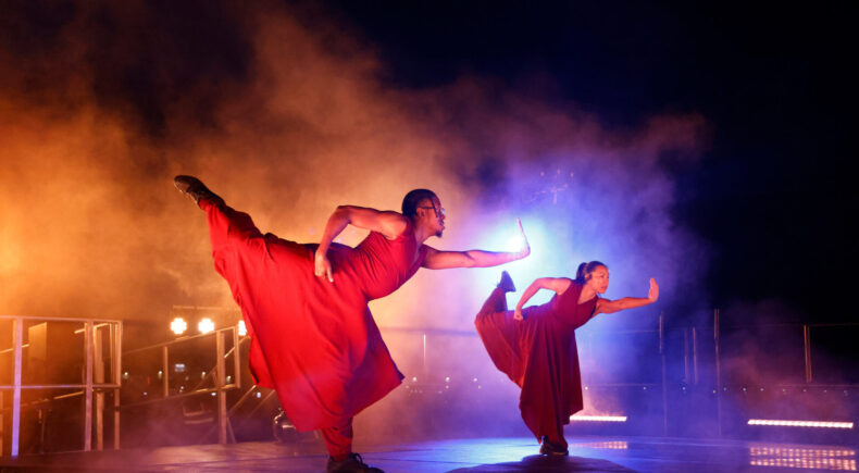Two dancers with outstretched arms and legs are light from behind. They wear baggy red clothing.
