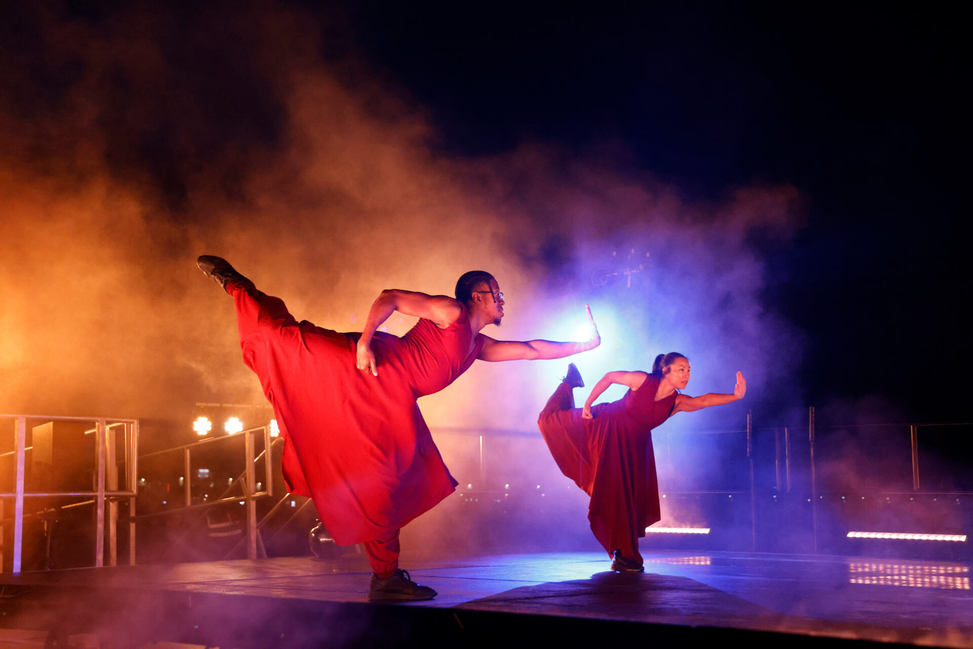 _U4A4913-Edit copy Lindsey Harris Two dancers with outstretched arms and legs are light from behind. They wear baggy red clothing.