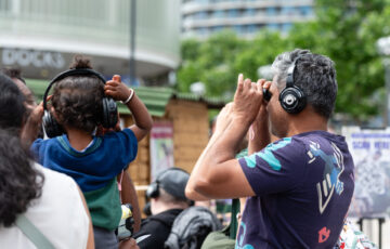 A man wearing headphones looks through some binoculars. A young boy also wears headphones
