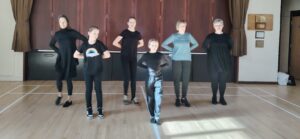 Young and older female dancers stand on a wooden floor with their arms behind their backs.