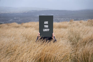 Hands holding a black card with white writing that says 'Are You Lost?', set in a wheat field