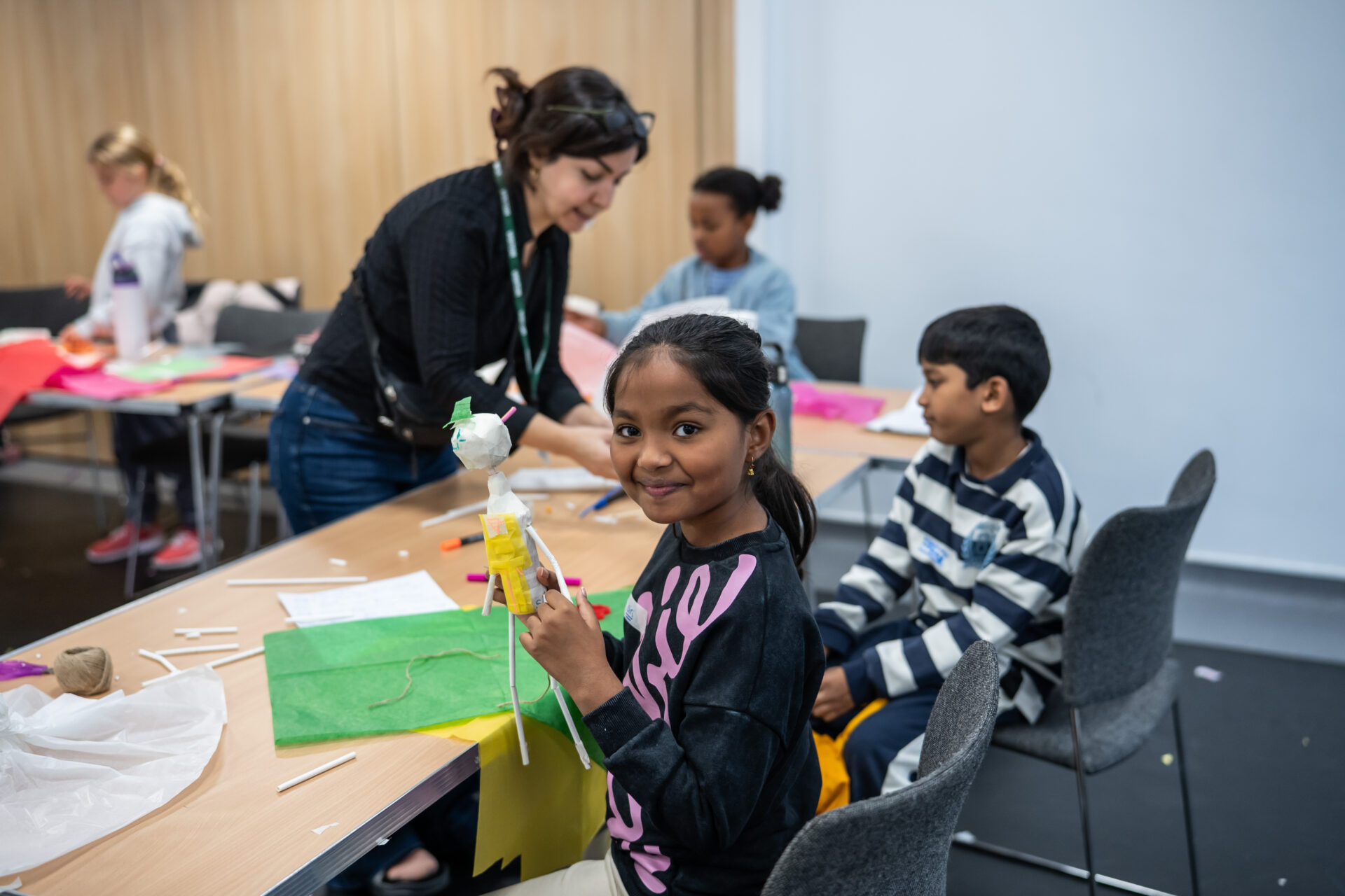 A young girl from the global majority, looks at the camera smiling. She is holding something in her hand. A woman helps the boy behind her.