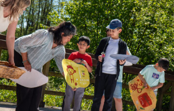 A group of young boys show their paintings to a woman of Indian heritage. There are trees in the background.