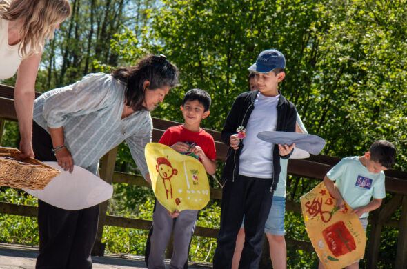 A group of young boys show their paintings to a woman of Indian heritage. There are trees in the background.
