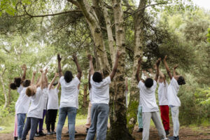A group of children are surrounding a tree in a forest, they are all wearing white tshirts and lifting their arms in the air