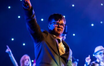 A boy stands on stage wearing a jacket and bow tie, pointing his finger in the air.