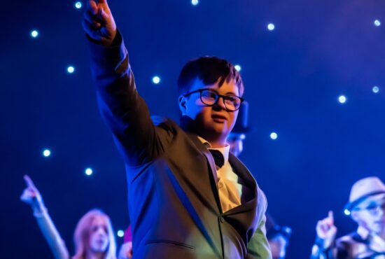 A boy stands on stage wearing a jacket and bow tie, pointing his finger in the air.