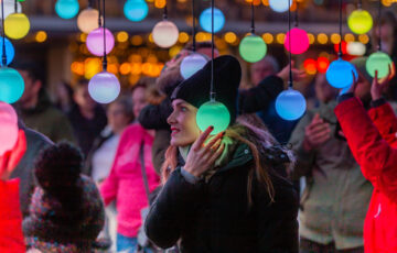 A white woman wearing a black coat and hat holds a glowing green orb to her ear. Other people are seen in the background.