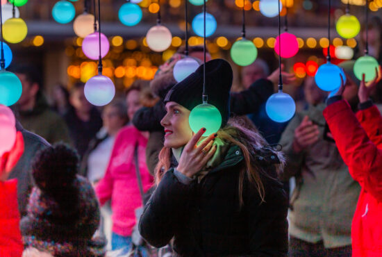 A white woman wearing a black coat and hat holds a glowing green orb to her ear. Other people are seen in the background.