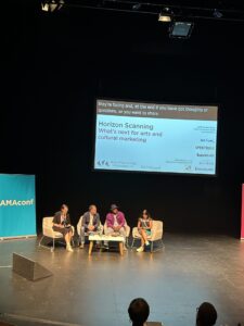 Four people sat on chairs on a flat floor stage. There is a screen behind them.