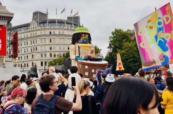 A crowd watch a large puppet, there are banners nearby
