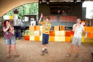 A young man holds one hand in the air in triumph. A woman and a man in summer clothing stand either side of him. There is a colourful stage behind them.