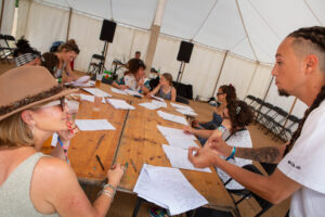 A man with dreadlocks talks to a women wearing a hat and sunglasses. There is a table and pieces of paper behind them, with other people sat at the table.