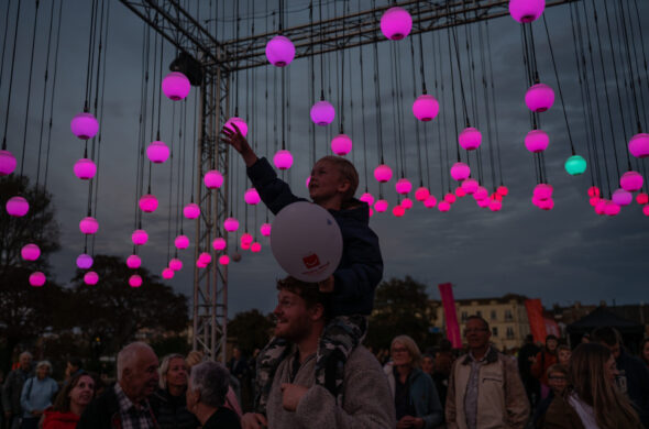 A crowd of people. A boy on a man's shoulders reaches out to touch one of many suspended pink orbs above them.