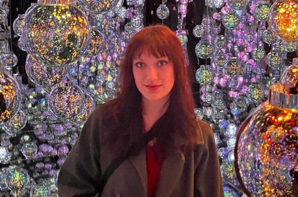 Young woman wearing a coat stands in front of lots of shining glass bulbs.