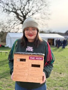 Young woman wearing glasses and a woolly hat hoplds a wooden shed-like model with text 'The Hide'